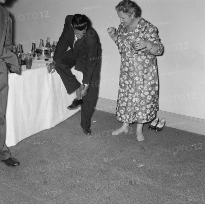 Removing shoes at a cocktail party. A suited man leans on a table for support as he removes his shoe at a Prudential Insurance cocktail party. He is encouraged by a middle-aged woman who is already barefoot and ready to dance. Kenya, 16 October 1957. Kenya, Eastern Africa, Africa.