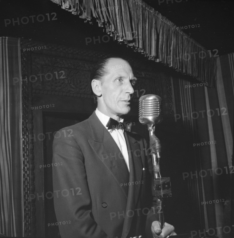 On stage at the Equator Club. A European man stands before a microphone on a small, curtained stage at the Equator Club. Related images suggest he is a singer with a band of African musicians. Nairobi, Kenya, 19 October 1957. Nairobi, Nairobi Area, Kenya, Eastern Africa, Africa.