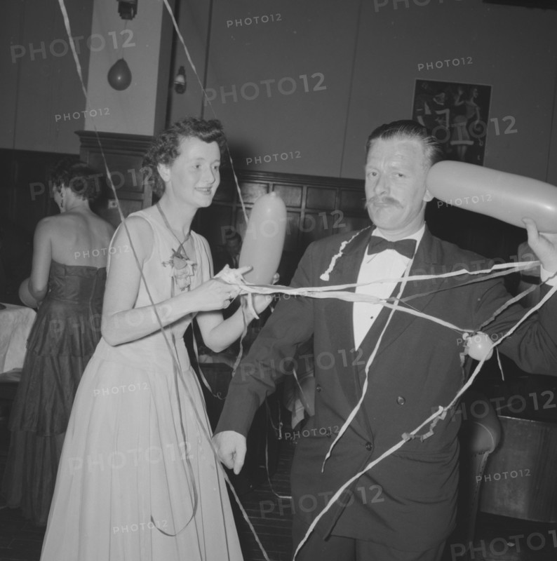 Single balloon salute. A middle-aged man at a Northern Counties dance, salutes the camera with a balloon as his partner looks on, amused. Kenya, 2 November 1957. Kenya, Eastern Africa, Africa.