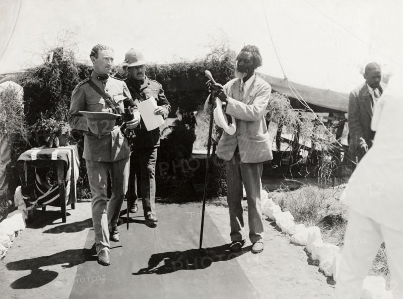 Entrance to a Presentation of Address. An African man, dressed in a Western-style suit, adjusts a long staff at the entrance to a Presentation of Address for the Duke and Duchess of Connaught. Arriving guests file past him along a red carpet. Gwelo, Southern Rhodesia (Gweru, Zimbabwe), 16 November 1910. Gweru, Midlands, Zimbabwe, Southern Africa, Africa.