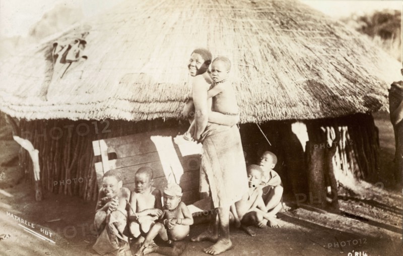 A Matabele family. A Matabele (Ndebele) woman turns to smile for the camera, a young child clinging to her back and five more sitting around her feet. An original caption comments: 'One man may have a doz(en) wives, hence the number of piccininas (children)'. Probably Southern Rhodesia (Zimbabwe), circa 1910. Zimbabwe, Southern Africa, Africa.