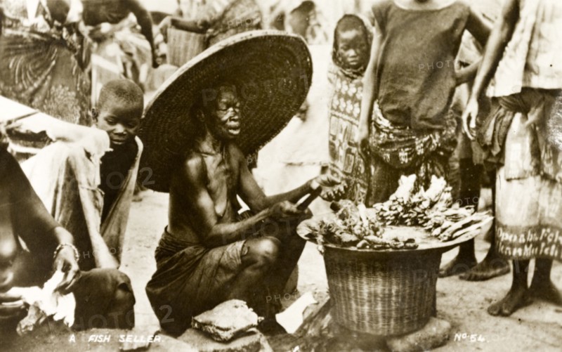 Fish seller, Gold Coast. An elderly woman dressed in a huge, wide brimmed hat, sells fish to passers by on a crowded street. Gold Coast (Ghana), circa 1910. Ghana, Western Africa, Africa.