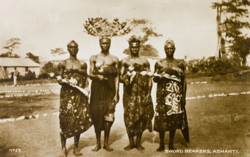 Asante swordbearers. Group portrait of four traditionally dressed Asante (Ashanti) swordbearers. One wears a feathered headdress and each holds a ceremonial sword. Gold Coast (Ghana), circa 1910. Ghana, Western Africa, Africa.