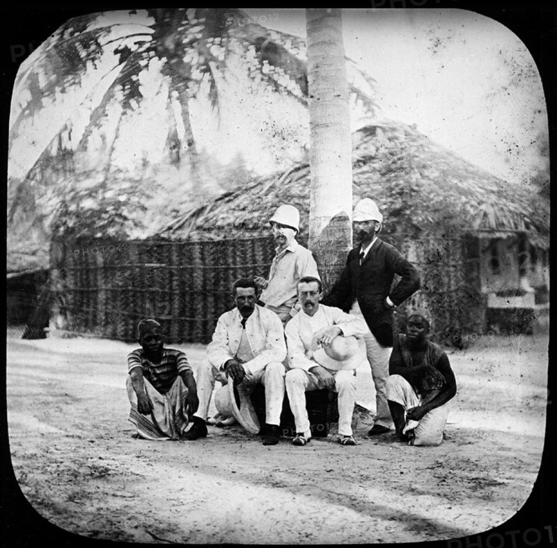 Consuls and explorers. Outdoors portrait of four European consuls and explorers, posing for the camera with their African servants. Eastern Africa, circa 1905., Eastern Africa, Africa.