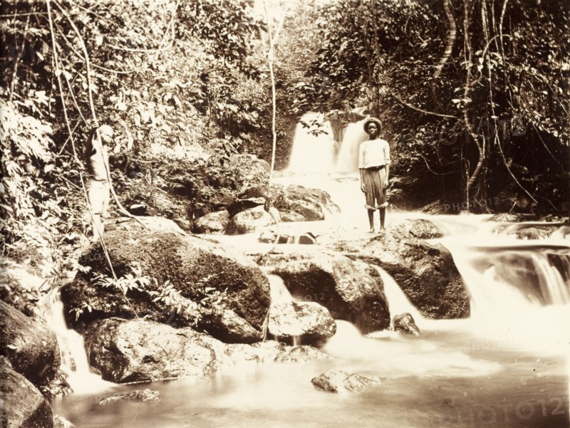 Jungle river in Bioko. Two young men stand on a rock formation straddling a jungle river. The slow shutter speed used by the photographer gives the water an ethereal quality as it cascades over the rocks in waterfalls. Bioko, Equatorial Guinea, circa 1930., Bioko Norte, Equatorial Guinea, Central Africa, Africa.