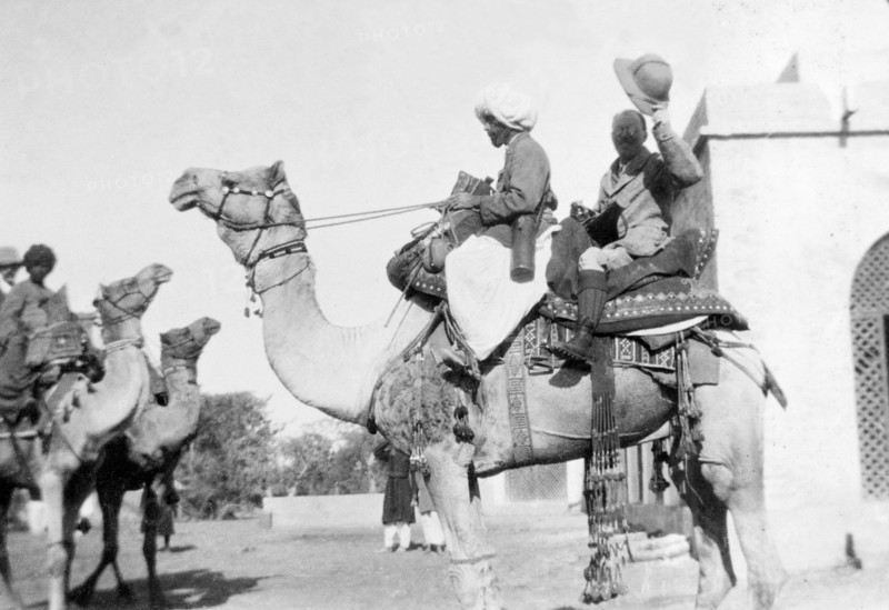 A hunting trip by camel. Sir Henry Staveley Lawrence, Collector of Karachi, raises his solatopi hat to the camera from the back of a camel. He sits behind an Indian 'shikari' (professional hunter), ready to embark on a hunting expedition to the countryside. Probably Sind, India (Sindh, Pakistan), December 1911., Sindh, Pakistan, Southern Asia, Asia.