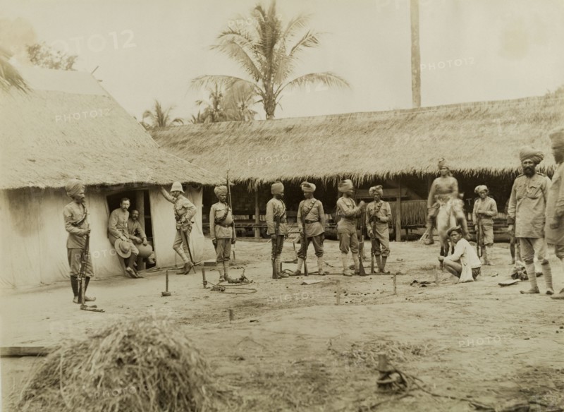 British and Indian soldiers in Burma (Myanmar). Armed British and Indian soldiers, possibly a mix of British Army and Military Police officers, mill about outside two thatched roof dwellings. A series of wooden stakes hammered into the ground appear to be fitted with restraints: perhaps a method of detaining captured prisoners. Sagaing, Burma (Myanmar), 1891., Sagaing, Burma (Myanmar), South East Asia, Asia.