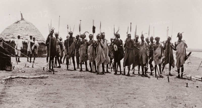 A group of Shilluk warriors. Portrait of a group of Shilluk warriors with their spears. The men stand closely together, dressed in simple robes with plumes of feathers in their hair. Taufikia, Sudan, circa 1910. Taufikia, Upper Nile, Sudan, Eastern Africa, Africa.