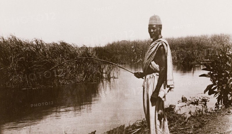 Fishing in the Jur River. A man stands fishing on the bank of the Jur River, holding a fishing rod in one hand whilst looking at the camera. Bahr-el-Ghazal region, Sudan, circa 1910., West Bahr el Ghazal, Sudan, Eastern Africa, Africa.