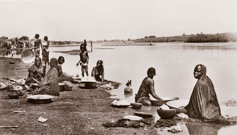 Washing clothes at the river bank, Sudan. Sudanese women and children wash their clothes on the banks of a river, using large, shallow bowls filled with river water. Waw, Sudan, circa 1910. Waw, West Bahr el Ghazal, Sudan, Eastern Africa, Africa.
