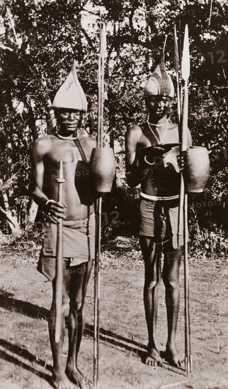 Portrait of two Sudanese men with spears. Portrait of two Sudanese men, identified by an original caption as 'sheikhs'. Semi-naked, they wear pointed hats and pose for the camera with their spears and hand shields. Bahr-el-Ghazal region, Sudan, circa 1910., West Bahr el Ghazal, Sudan, Eastern Africa, Africa.