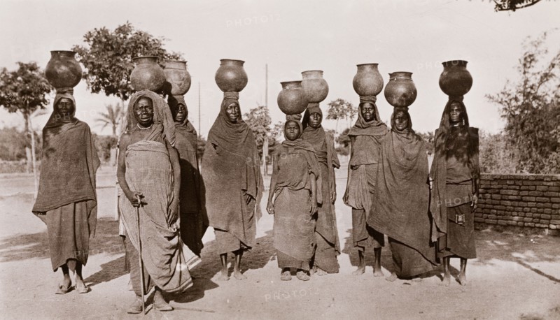 Sudanese women carrying water. A group of Sudanese women, dressed in long robes and headscarves, walk along a road carrying water in ceramic pots on their heads. Sudan, circa 1910. Sudan, Eastern Africa, Africa.