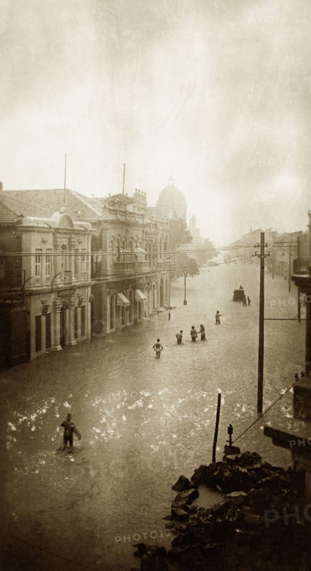 Flooded street in Karachi. A small band of people wades towards a partially submerged vehicle along a flooded street in Karachi. The water is knee-deep, almost reaching the ground-floor windows of the surrounding buildings. Karachi, India (Pakistan), circa 1910. Karachi, Sindh, Pakistan, Southern Asia, Asia.