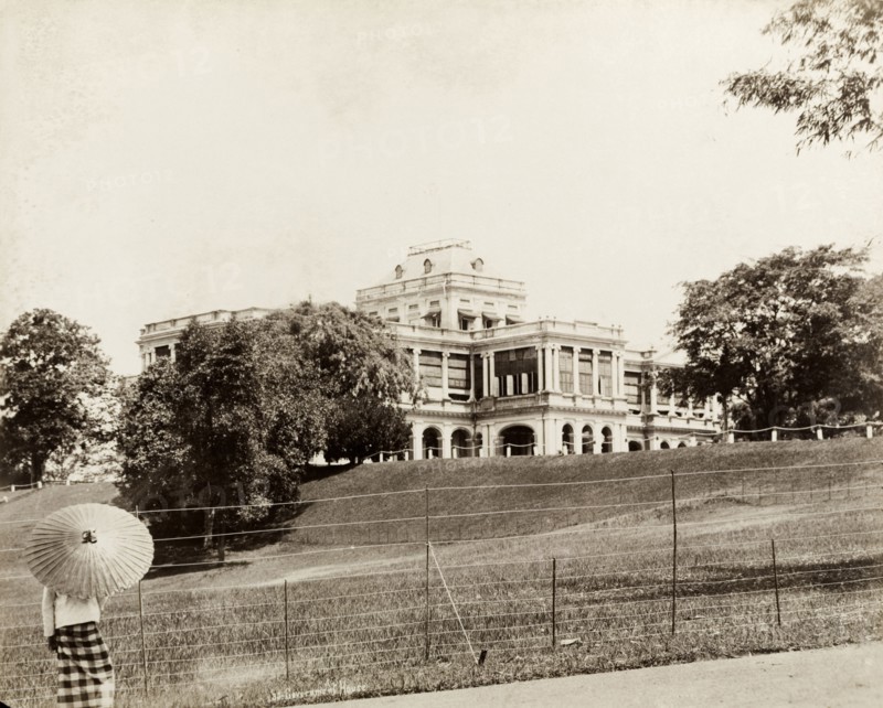 Government House, Singapore. Government House stands on a rise in landscaped gardens behind a wire fence. A person in local dress holding an Asian umbrella stands with their back to the camera in the left foreground. Singapore, Straits Settlements (Singapore), circa 1895. Singapore, Central (Singapore), Singapore, South East Asia, Asia.