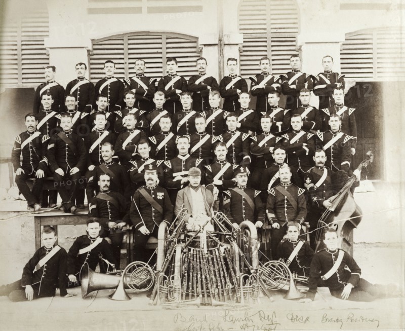 British Army military band with instruments. A British Army military band pose for a group portrait, seated in rows behind their musical instruments, which include clarinets, brass horns and a doube bass. The central figure wears a casual jacket and cap and holds what appears to be a squash racquet. Southern Asia, circa 1895., Southern Asia, Asia.