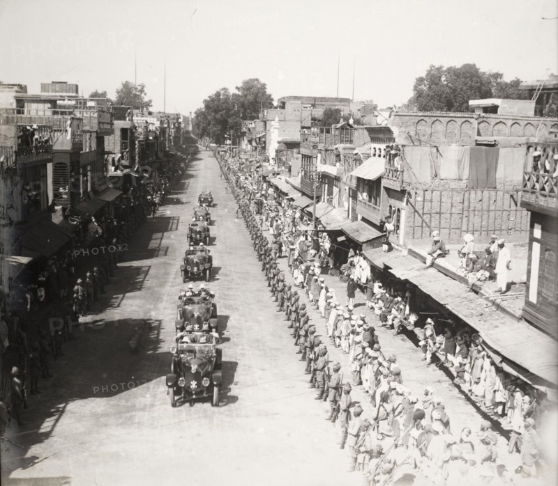 Indian Army officers salute a procession of cars. Hundreds of Indian Army officers, and crowds of civilians, line a city street as a procession of chauffeur-driven cars passes by. Each vehicle contains several British men in uniform, probably government or military officials on their way to a formal engagement. Probably northern India, circa 1923. India, Southern Asia, Asia.