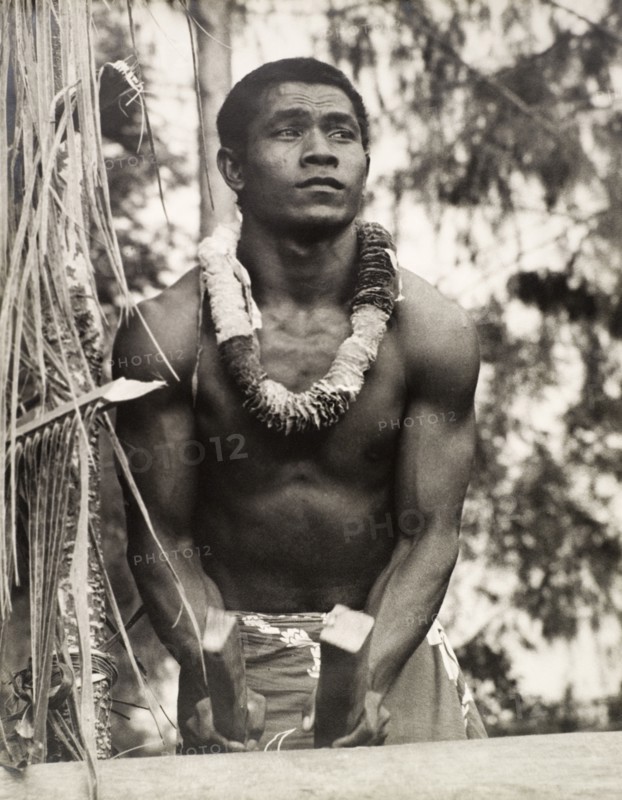 A secondary school student, Solomon Islands. A student of the King George VI School in Auki, drums using two roughly chopped pieces of wood as drumsticks. He is naked from the waist up and wears a floral garland around his neck. Auki, Malaita Island, Solomon Islands, circa 1960. Auki, Malaita, Solomon Islands, Pacific Ocean, Oceania.