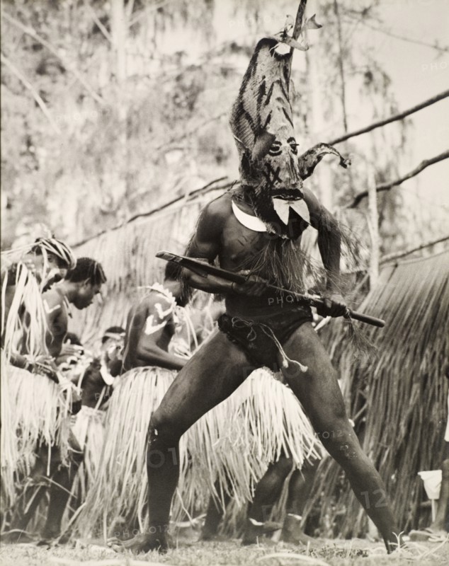 Student performing in traditional costume, Solomon Islands. A student of the King George VI School in Auki, wears traditional costume for a performance. He is dressed in an elaborate, painted mask, which covers his head, and holds a short spear with both hands. Auki, Malaita Island, Solomon Islands, circa 1960. Auki, Malaita, Solomon Islands, Pacific Ocean, Oceania.