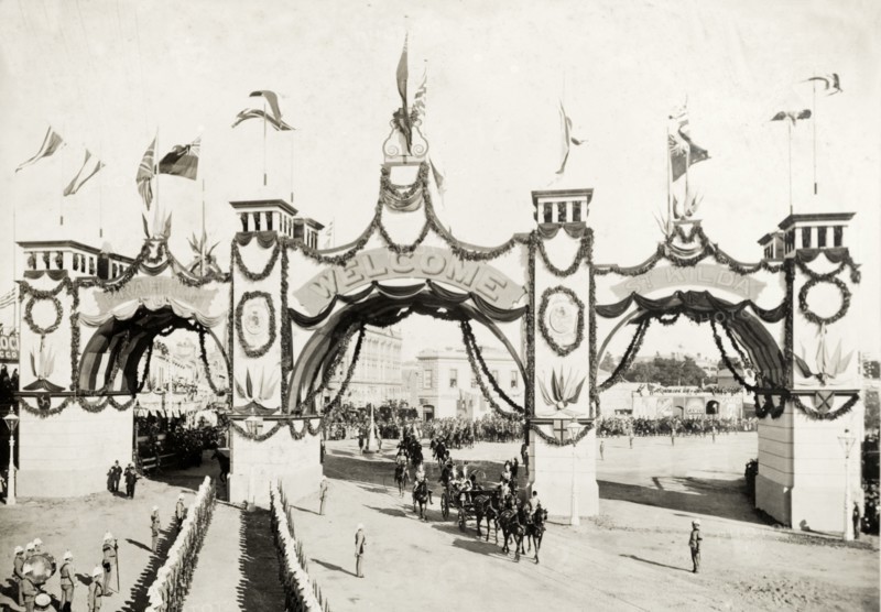 Welcome arch for the royal visit, Melbourne. A welcome arch, decorated with flags and garlands, spans a street in honour of the Duke and Duchess of Cornwall and York (later King George V and Queen Mary) who had travelled from England to open the first Commonwealth Parliament of Australia. A horse-drawn carriage passing beneath receives a military salute, suggesting its passengers are of some importance. Melbourne, Australia, May 1901. Melbourne, Victoria, Australia, Australia, Oceania.