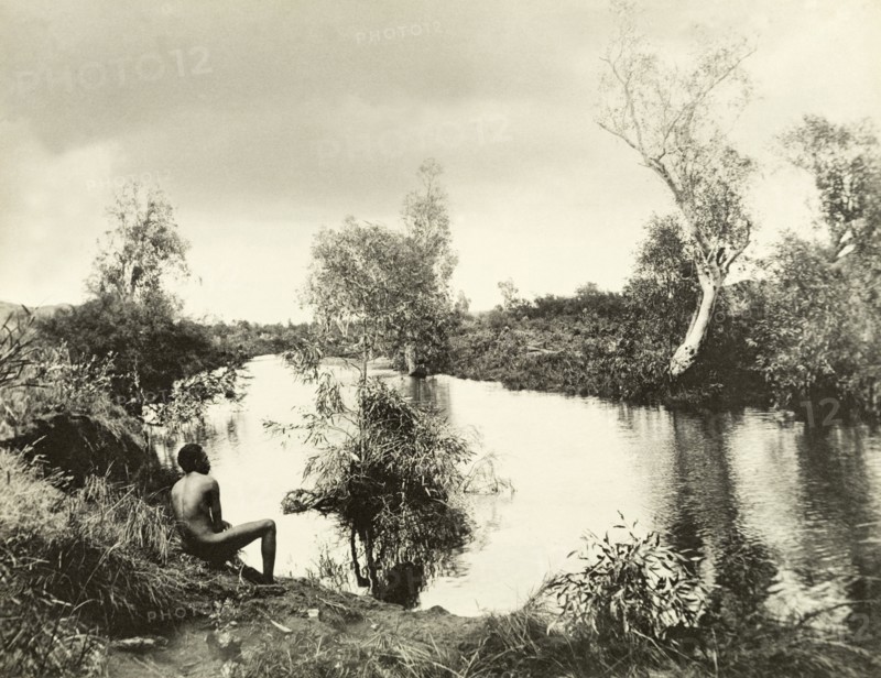 An aborigine on the banks of a backwater. An aborigine sits with his back to the camera on the banks of a peaceful backwater in the outback. Western Australia, Australia, circa 1901., West Australia, Australia, Australia, Oceania.