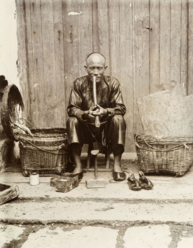 An itinerant shoemaker, Hong Kong. An itinerant shoemaker sits on a stool, smoking a long pipe at his street stall in Hong Kong. A shoe mould, a box of tools and a pair of sandals are positioned in front of him. Hong Kong, China, circa 1903. Hong Kong, Hong Kong, China, People's Republic of, Eastern Asia, Asia.