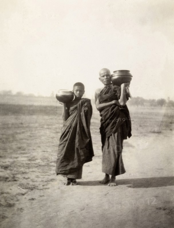 Pot carriers in Mandalay. A Burmese man and a boy dressed in long robes carry pots on their shoulders along a dusty road at Fort Dufferin. Mandalay, Burma (Myanmar), circa February 1927. Mandalay, Mandalay, Burma (Myanmar), South East Asia, Asia.
