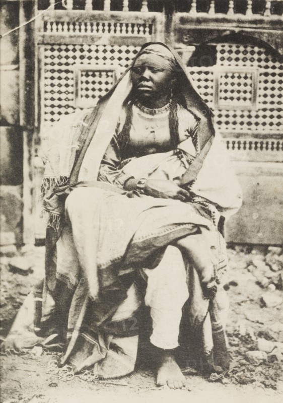 Portrait of an African woman in Egypt. Portrait of an African woman, sitting cross-legged on a chair in front of a decorative window screen. She wears a robe and head scarf, and gazes off to the left of the camera. Port Said, Egypt, circa 1920. Port Said, Port Said, Egypt, Northern Africa, Africa.