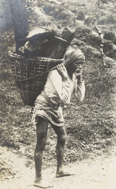 Transporting firewood in the hills. Portrait of an elderly man carrying a basket full of firewood in the hills. Hunched over and barefoot, he bears the load with the aid of a strap attached to his head. Probably Nepal, circa 1922. Nepal, Southern Asia, Asia.