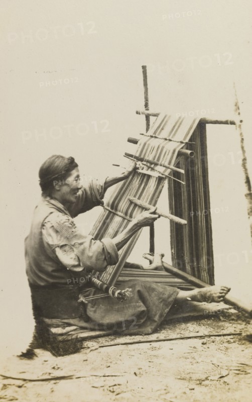Bhatia woman weaving cloth. An elderly Bhatia woman sits on the ground, operating a frame loom by hand to weave a length of patterned cloth. India, circa 1922. India, Southern Asia, Asia.