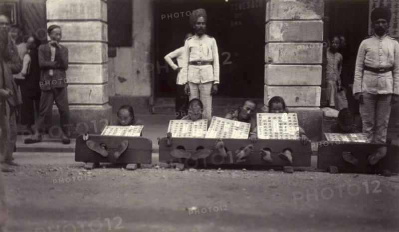 Petty criminals in the stocks, Hong Kong. A group of petty criminals sit with their ankles restrained in stocks, guarded by a pair Indian officers from the Hong Kong Police Force. Each wears a board strung around the neck, describing in detail the offense committed. Hong Kong, China, circa 1905. Hong Kong, Hong Kong, China, People's Republic of, Eastern Asia, Asia.