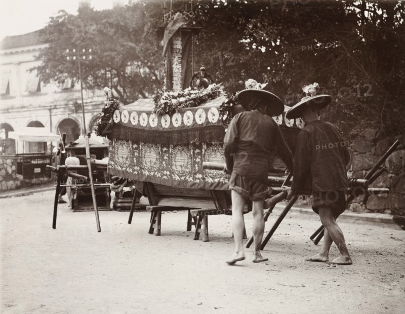 Pallbearers at a Chinese funeral. Two pallbearers prepare to carry a coffin on a wooden bier during a Chinese funeral ceremony. The coffin is draped in fine cloth and decorated with wreaths of flowers, indicating that the deceased had been wealthy. China, circa 1905. China, People's Republic of, Eastern Asia, Asia.