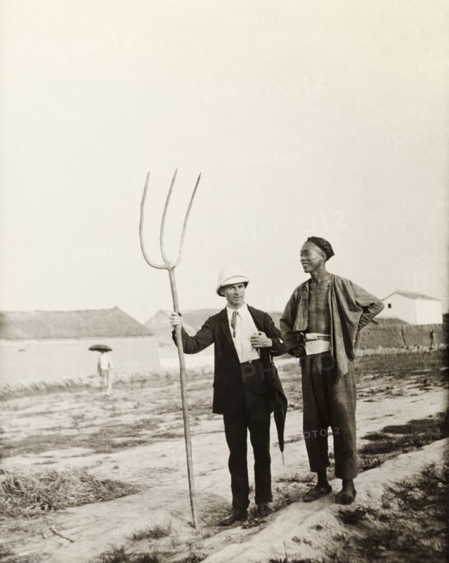 European man with Chinese farming tool. A European man, dressed in a suit and solatopi hat, holds a large, three-pronged fork as he chats to a Chinese farm labourer. Northern China, circa 1920. China, People's Republic of, Eastern Asia, Asia.