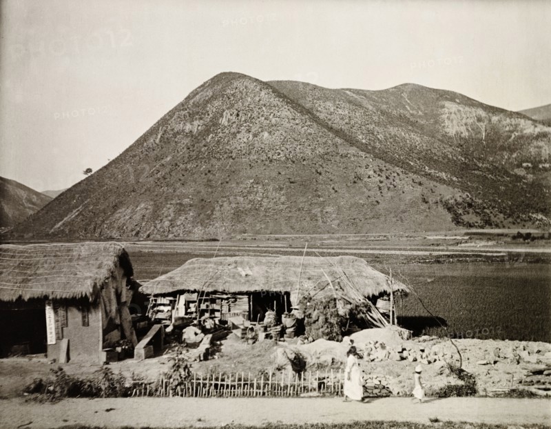 Dwelling in the mountains, Korea. A small dwelling with a thatched roof sits at the edge of a flat plain against a mountainous backdrop. Korea (South Korea), circa 1920. South Korea, Eastern Asia, Asia.