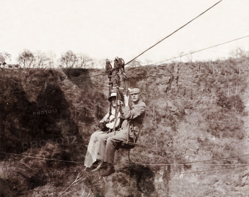 Lady Lawley and Arthur Rhodes cross the Zambezi gorge. Lady Annie Lawley, the wife of Sir Arthur Lawley, Lieutenant-Governor of the Transvaal, and Arthur Rhodes (brother of Cecil John Rhodes) travel across the Zambezi gorge at Victoria Falls on a chair lift attached to a cable system. Matabeleland, Rhodesia (Matabeleland North, Zimbabwe), circa 1904., Matabeleland North, Zimbabwe, Southern Africa, Africa.