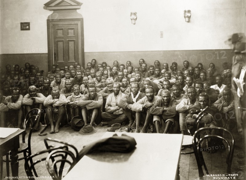 A Matabele indaba with Captain Lawley. Matabele indunas (chiefs) sit on the floor of the Stock Exchange Hall with their arms wrapped around their knees. The group were attending an indaba (council) headed by Captain Arthur Lawley, Administrator of Matabeleland and Mashonaland, and the Chief Native Commissioner to discuss arrangements for administering Matabeleland. Bulawayo, Rhodesia (Zimbabwe), 1897. Bulawayo, Matabeleland North, Zimbabwe, Southern Africa, Africa.