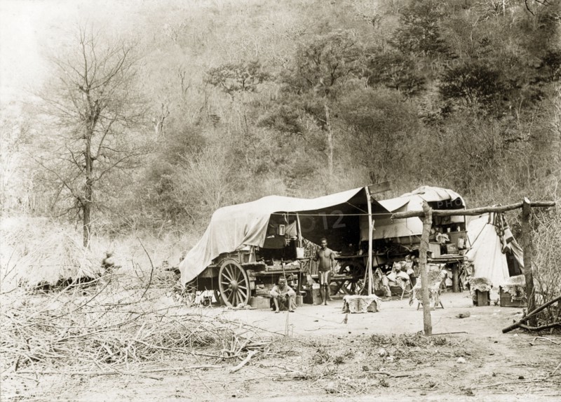Camp site in the bush. Tents are pitched beside wagons covered with canvas at a camp site in the bush. Two African guides accompany a European man who reclines on a chair lined with animal skin. Near Wankie, Rhodesia (Hwange, Zimbabwe), circa 1898. Hwange, Matabeleland North, Zimbabwe, Southern Africa, Africa.