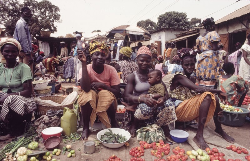 Female street traders at Banjul market. Female traders sell fruit and vegetables at a bustling outdoor market in Banjul. One of the women balances a baby on her knee, whilst another holds up a smoked fish for the camera. Banjul, Gambia, circa 1985. Banjul, Banjul City, Gambia, Western Africa, Africa.