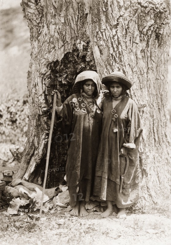 Two women from Kashmir. Portrait of two women from Kashmir, standing at the foot of a giant tree. Both women have their heads covered and wear long robes decorated with hoop brooches. One leans on a staff, whilst the other holds a sickle. Jammu and Kashmir State, India, circa 1925., Jammu and Kashmir, India, Southern Asia, Asia.