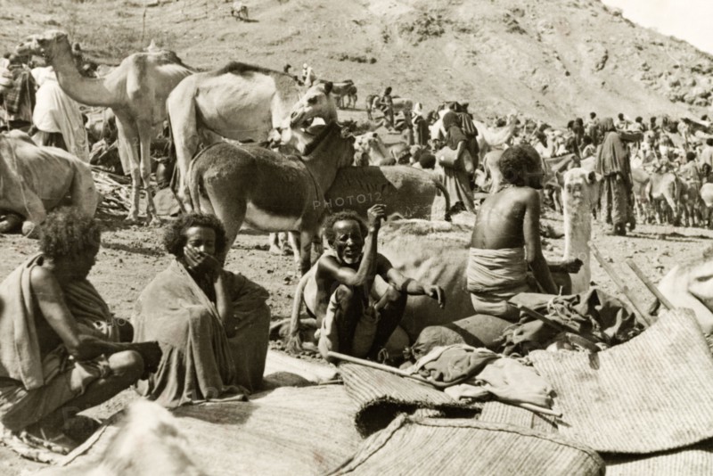 Eritrean men at a camel market. Four Eritrean men sit beside a heap of woven mats at a bustling camel market. Eritrea, 1943. Eritrea, Eastern Africa, Africa.
