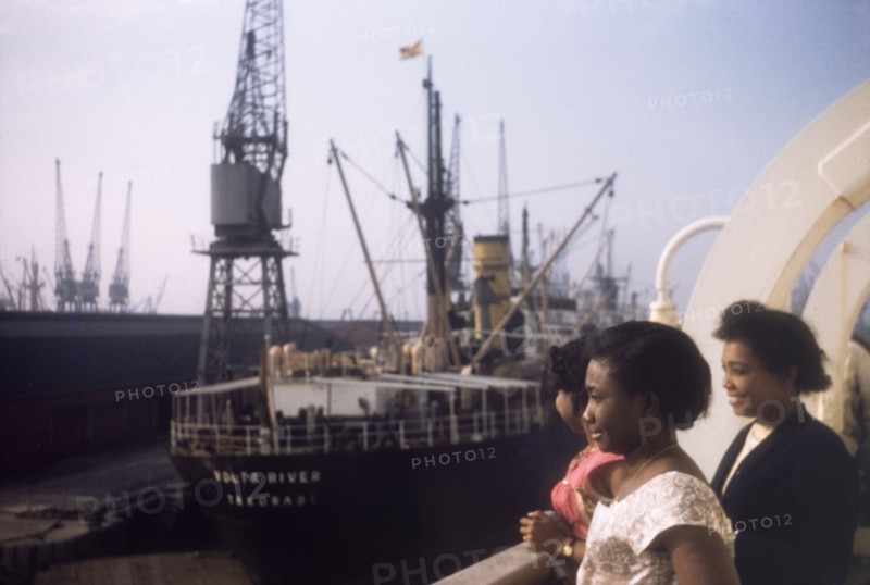 Shipping at Tilbury Docks. Three West African women gaze over the side of a ship moored at Tilbury Docks on the River Thames. A large steamship, 'River Volta', sits in harbour in the distance. Tilbury, England, September 1958. Tilbury, Essex, England (United Kingdom), Western Europe, Europe .