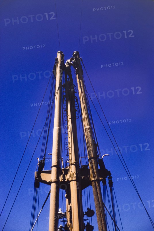Sailor on the mast of M.V. Obuasi. A sailor stands at the top of a ladder on the stern mast of a cargo ship, M.V. Obuasi, adjusting or repairing the rigging. Ghana, circa 1960. Ghana, Western Africa, Africa.