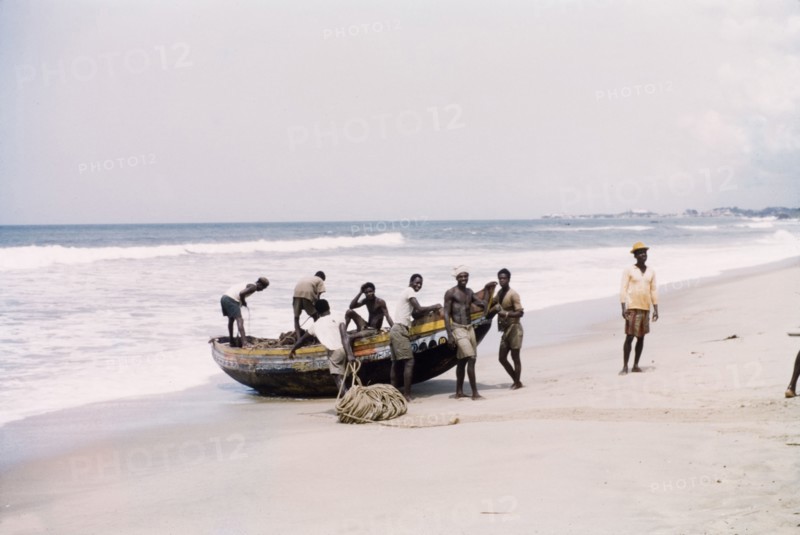 Fishing boat at Cape Coast. A group of men stand around a small fishing boat that is grounded on a beach. Near Cape Coast, Ghana, circa 1958. Cape Coast, Central (Ghana), Ghana, Western Africa, Africa.
