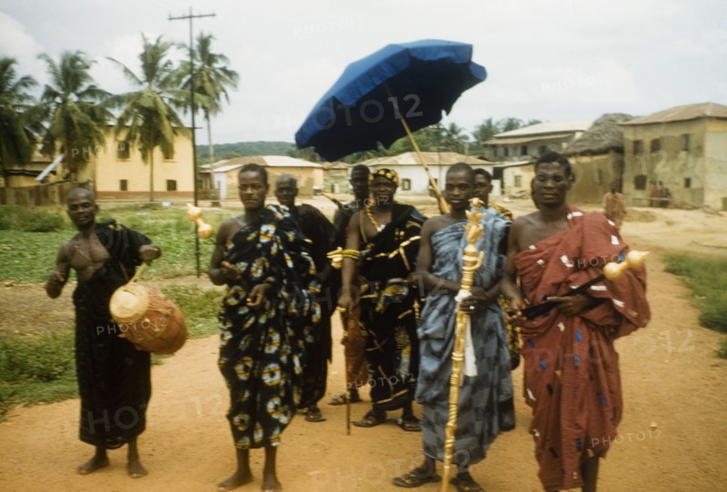 An Ghanaian chief and his retinue. A Ghanaian chief and his retinue process along a road in Anomabu. Members of the entourage wear traditional dress and carry various items including a golden staff, a ceremonial umbrella and a drum. Anomabu, Ghana, January 1958. Anomabu, Central (Ghana), Ghana, Western Africa, Africa.