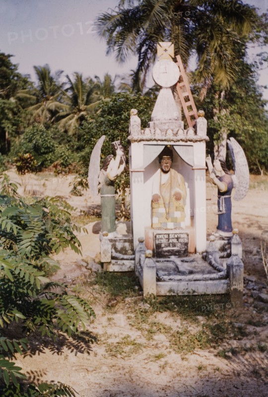 Christian grave on the Winneba-Shedru Road. A Christian grave or burial monument is marked with a stone cross on the Winneba-Shedru Road. The elaborately sculpted and painted headstone shows a seated dignitary wearing kente cloth, flanked by angels who point to heaven. It is topped with a conical tower featuring a clock, a Maltese cross and a ladder. Central Region, Ghana, circa 1960., Central (Ghana), Ghana, Western Africa, Africa.