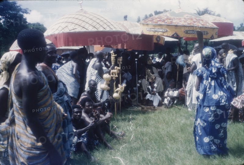 Mate Kole II at a Ngmayem festival. Mate Kole II (1910-1990), the Paramount Chief or Konor of Manya Krobo, sits beneath ceremonial umbrellas with his retinue at an annual Ngmayem harvest festival. He is attended by his 'okyeames' (linguists), each of whom carries a 'poma' (linguist's staff) as a badge of office. The child with the painted face and elaborate headdress, seated at Mate Kole II's feet, represents the Konor's 'soul'. Odumasi, Ghana, circa 1960. Odumasi, East (Ghana), Ghana, Western Africa, Africa.