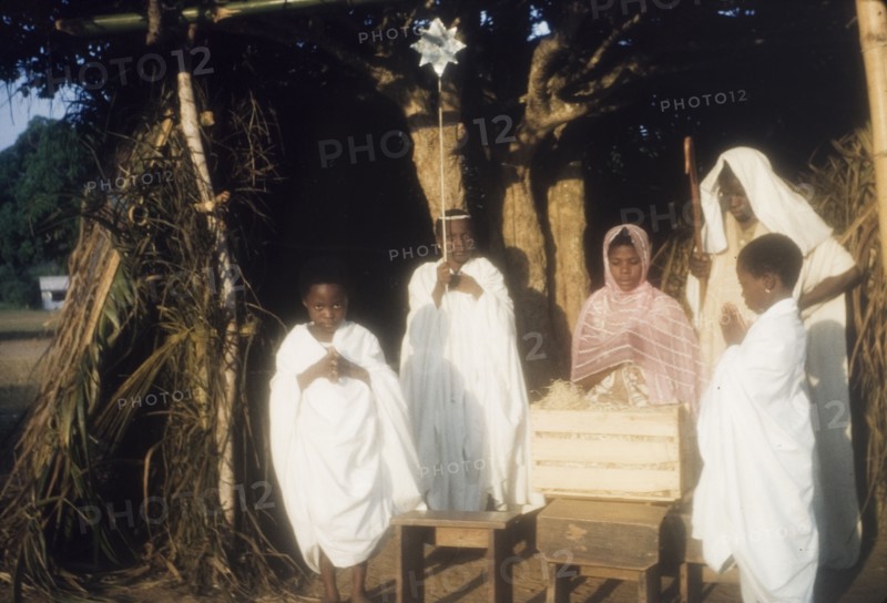 Ghanaian children perform a nativity play. Children in fancy dress perform a nativity play outdoors. Dressed as Mary, Joseph and the angels, they stand with their hands pressed together in prayer beside a straw-filled crate representing the crib of baby Jesus. Ghana, circa 1960. Ghana, Western Africa, Africa.