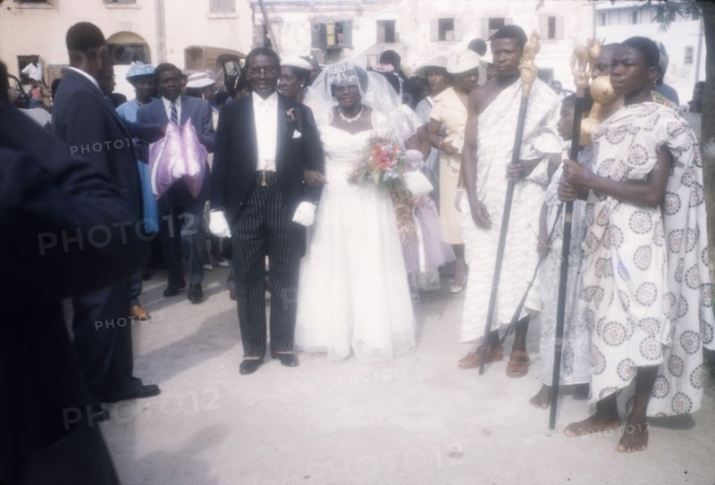 Christian wedding in Ghana. A Ghanaian bride arrives for her wedding accompanied by her father who wears a formal suit. Two men in traditional Ghanaian dress stand nearby holding ceremonial staffs. Ghana, 23 April 1960. Ghana, Western Africa, Africa.
