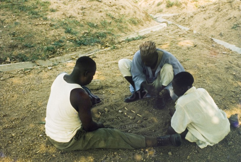 Playing a mancala game, Nigeria. Two men sit on the ground, playing a mancala game in the sand while a young boy looks on. Enugu, Nigeria, circa 1958. Enugu, Anambra, Nigeria, Western Africa, Africa.