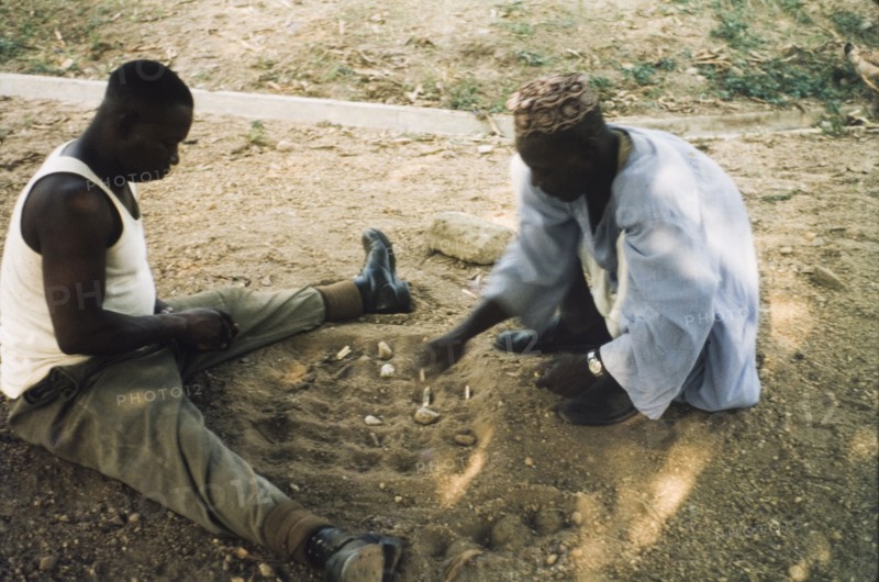 Playing a mancala game, Nigeria. Two men sit on the ground, playing a mancala game in the sand. Enugu, Nigeria, circa 1958. Enugu, Anambra, Nigeria, Western Africa, Africa.
