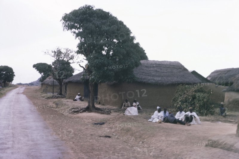 A Hausa village. Inhabitants of a Hausa village sit on the ground outside several square, mud-walled huts with thatched roofs. Nigeria, October 1963. Nigeria, Western Africa, Africa.